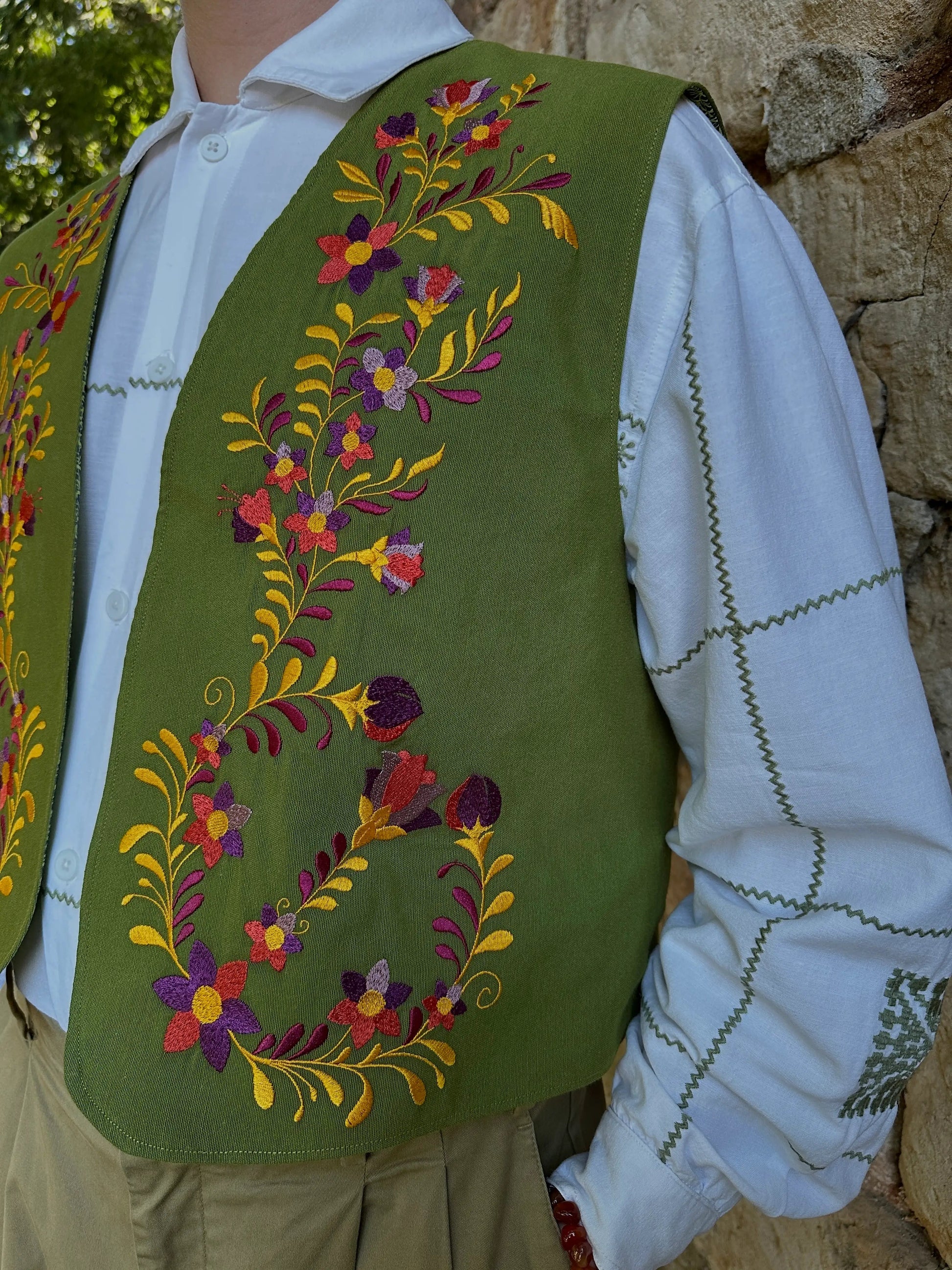 Person wearing a green embroidered vest over a white shirt against a stone wall.