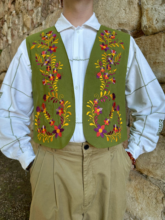 Person wearing a green embroidered vest over a white shirt with a stone wall background