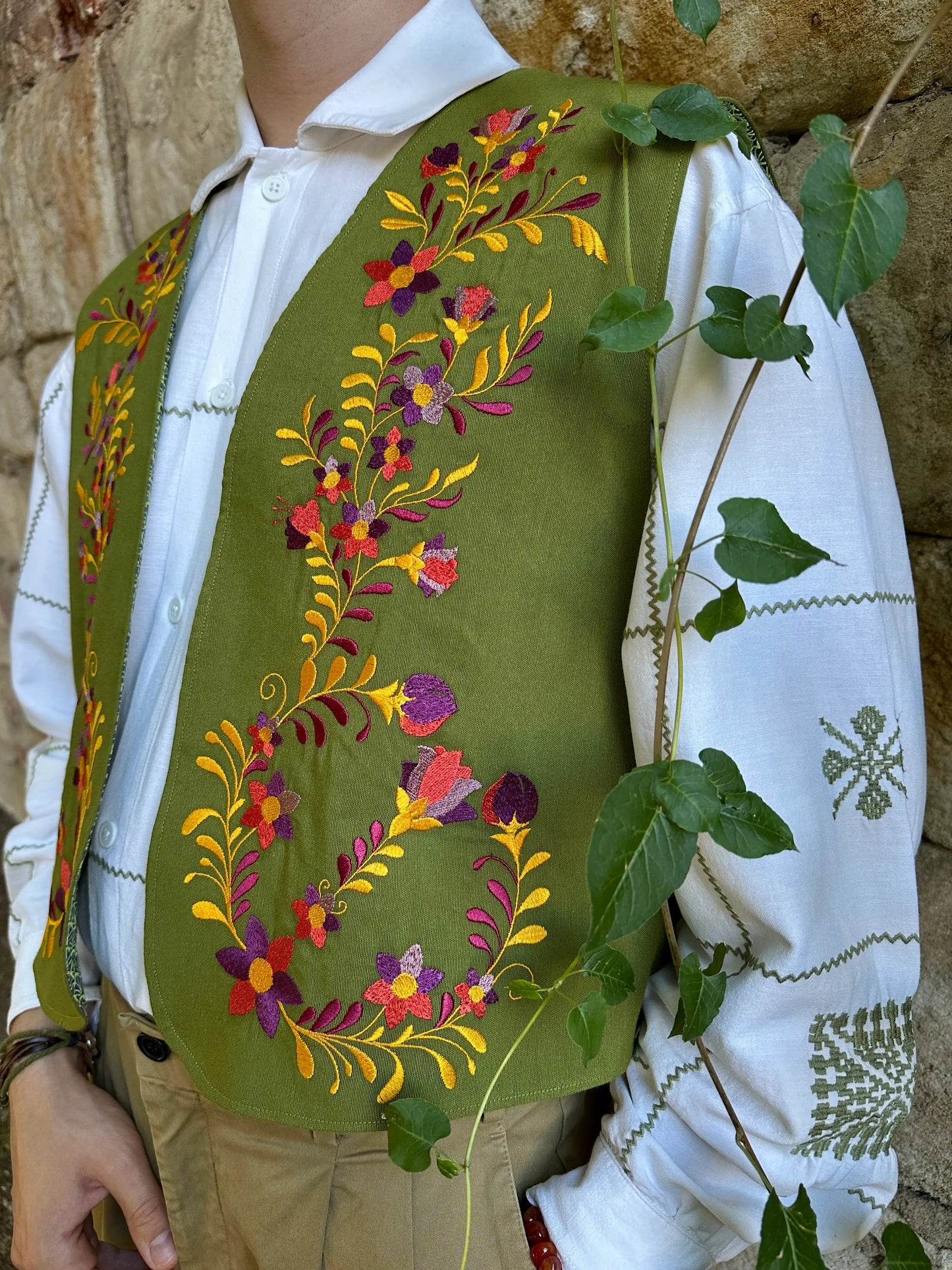 Green embroidered vest over a white shirt with floral patterns, against a stone wall background.