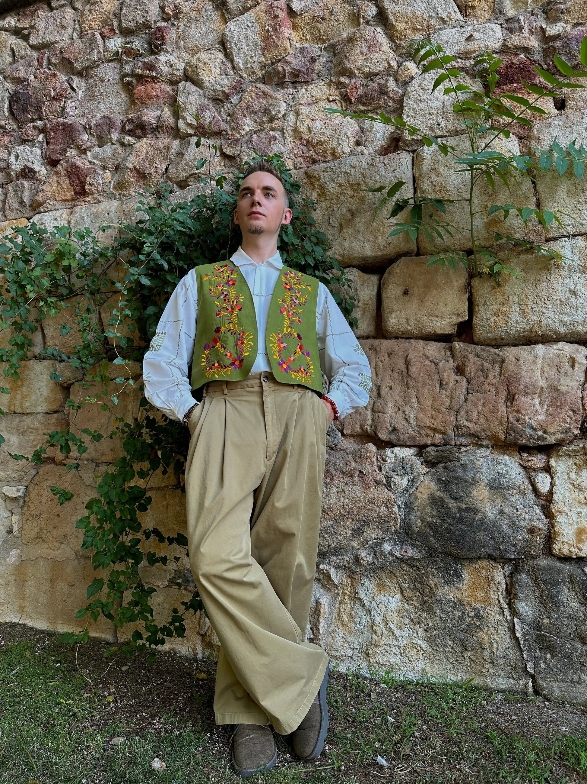 Man in traditional attire standing against a stone wall with greenery.
