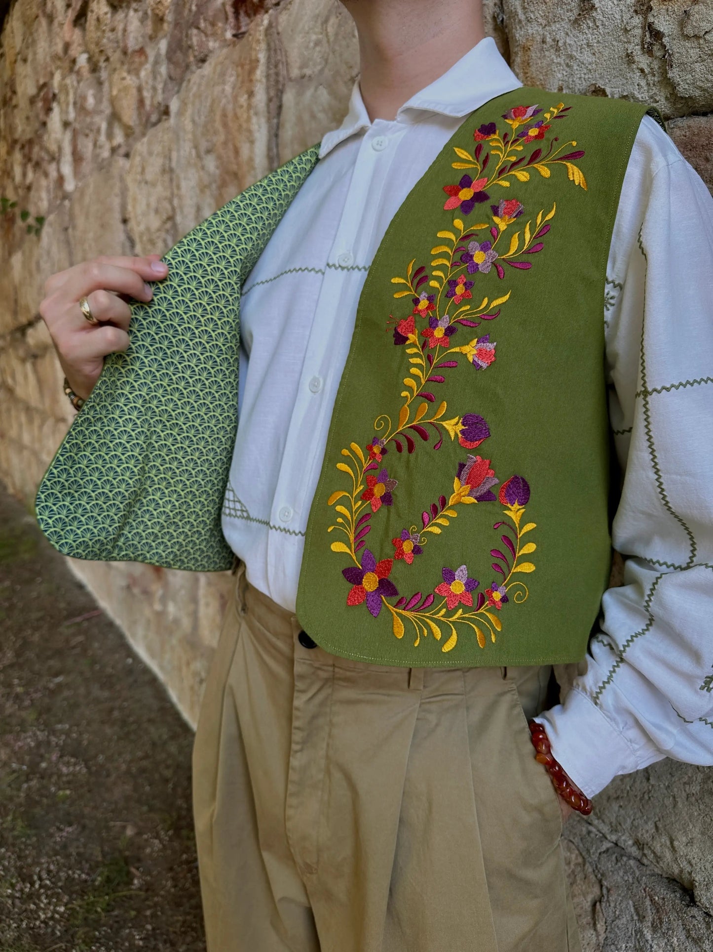Person wearing a green embroidered vest over a white shirt with beige pants, against a stone wall background.
