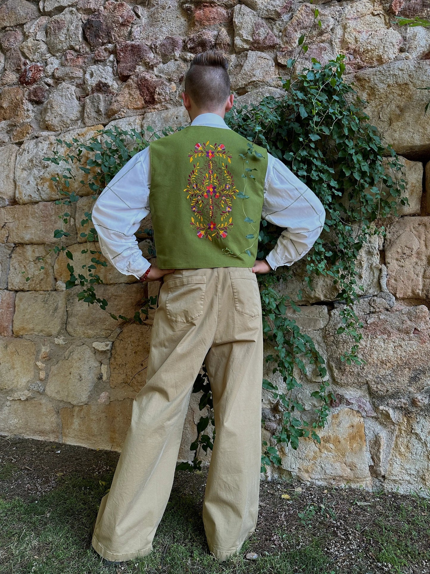 Person wearing a green embroidered vest and beige pants standing against a stone wall with greenery.