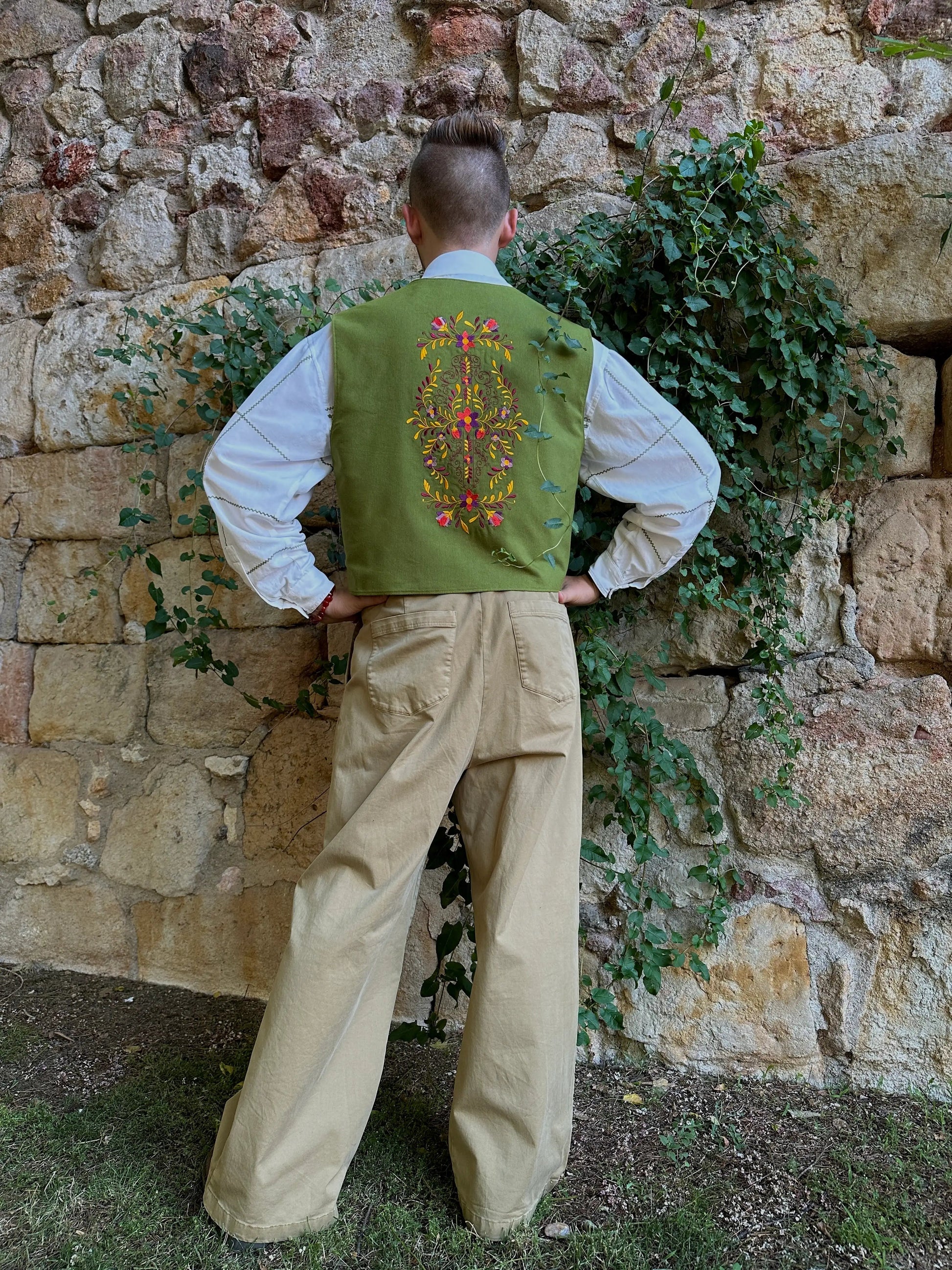 Person wearing a green embroidered vest and beige pants standing against a stone wall with greenery.