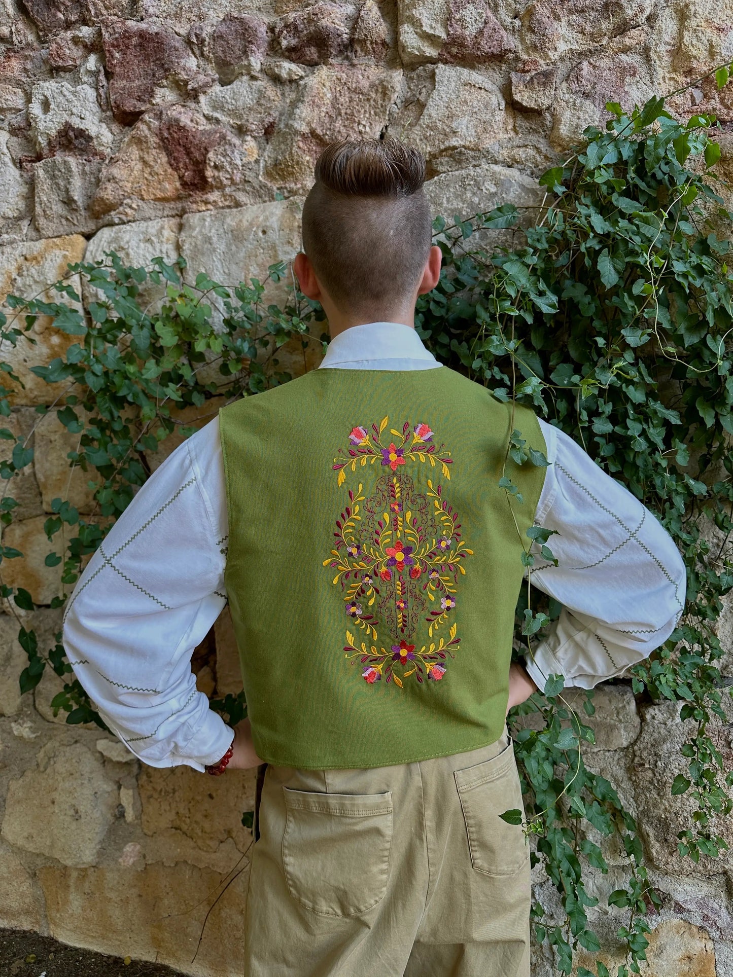 Person wearing a green embroidered vest standing against a stone wall with greenery.