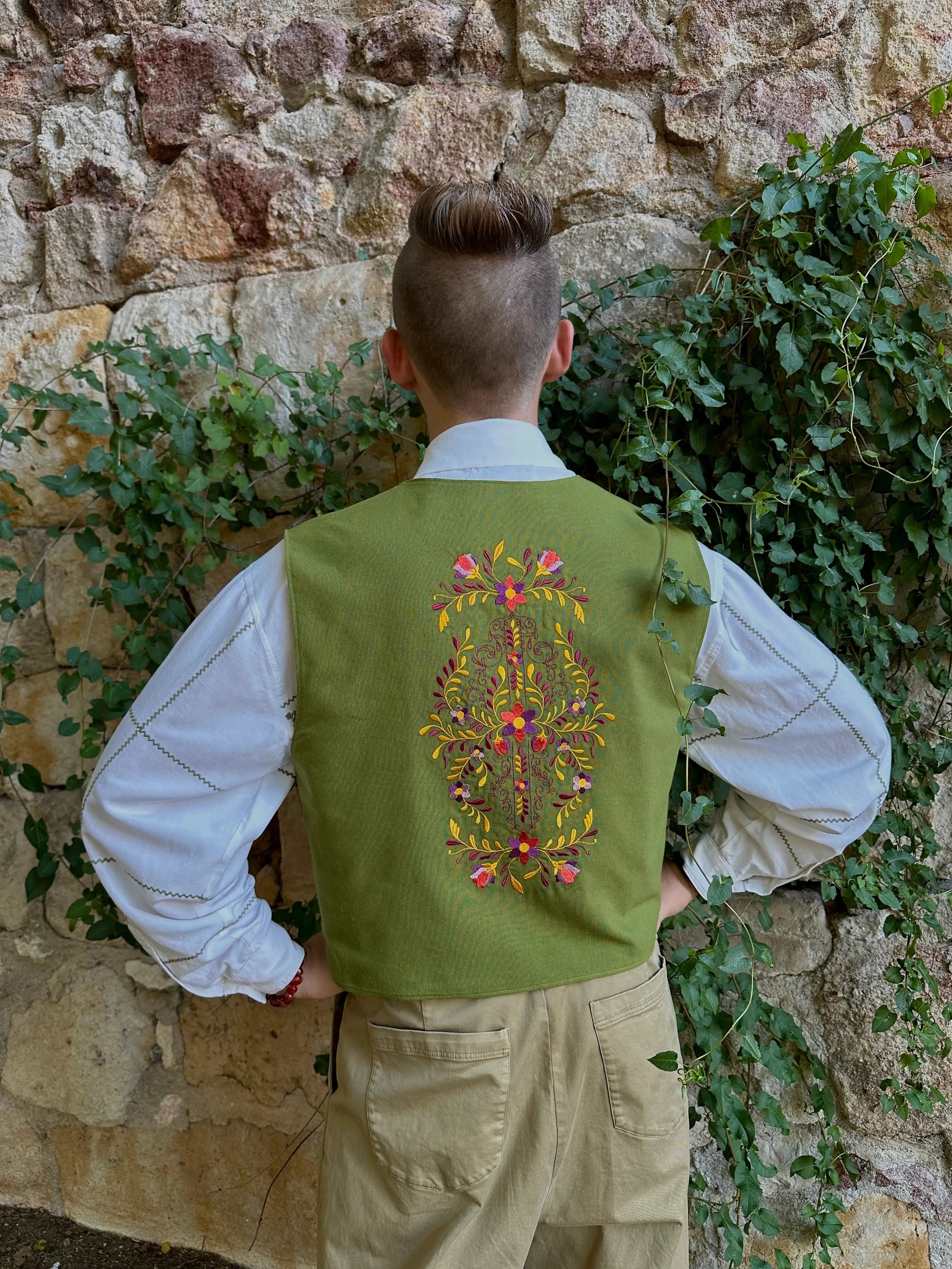 Person wearing a green embroidered vest standing against a stone wall with greenery.