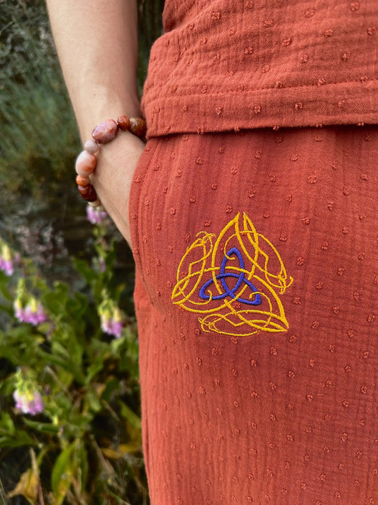 Person wearing a rust-colored garment with a Celtic knot design, holding a beaded bracelet against a natural background.