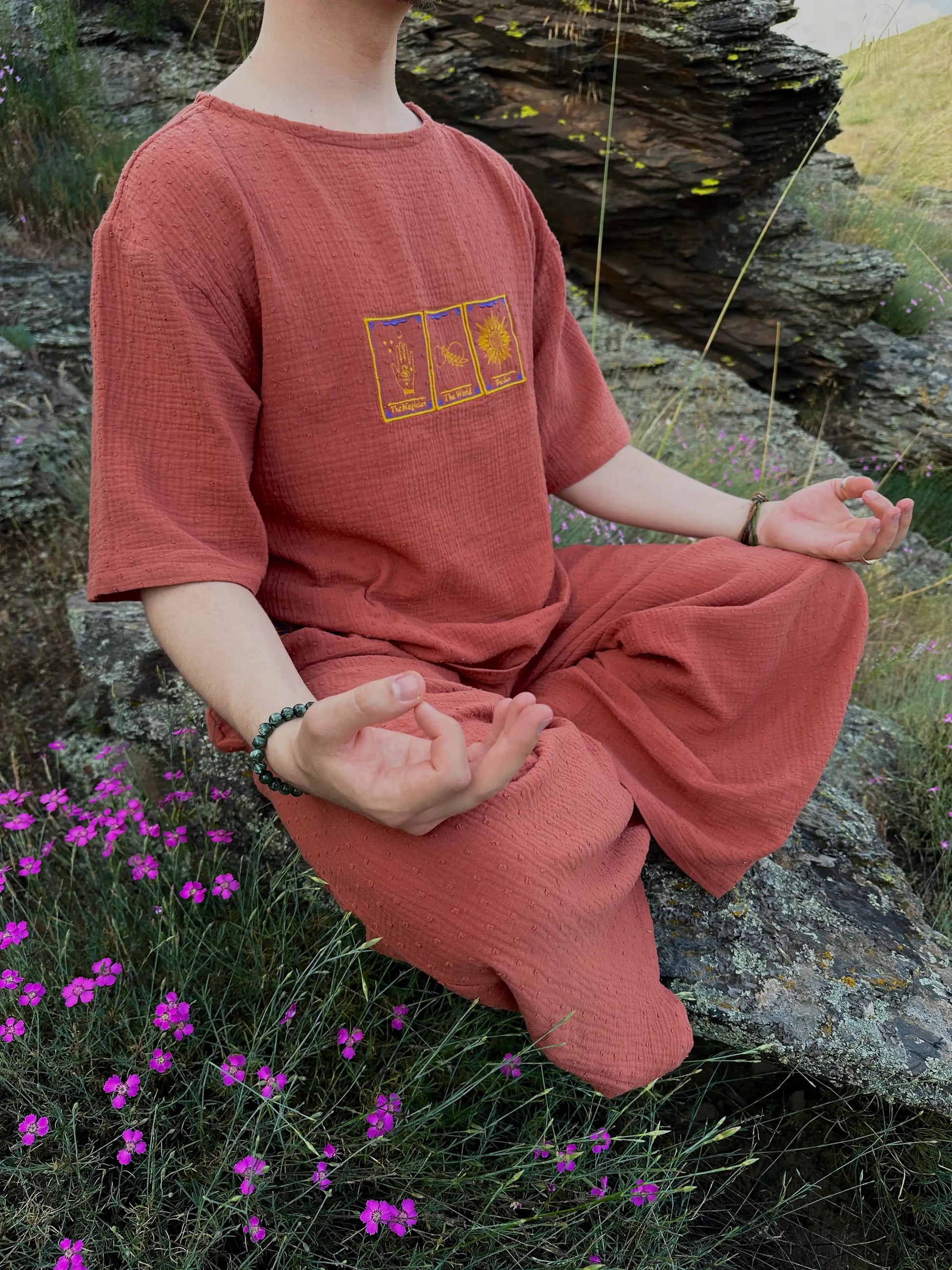 Person in a red outfit meditating outdoors with purple flowers and rocks in the background
