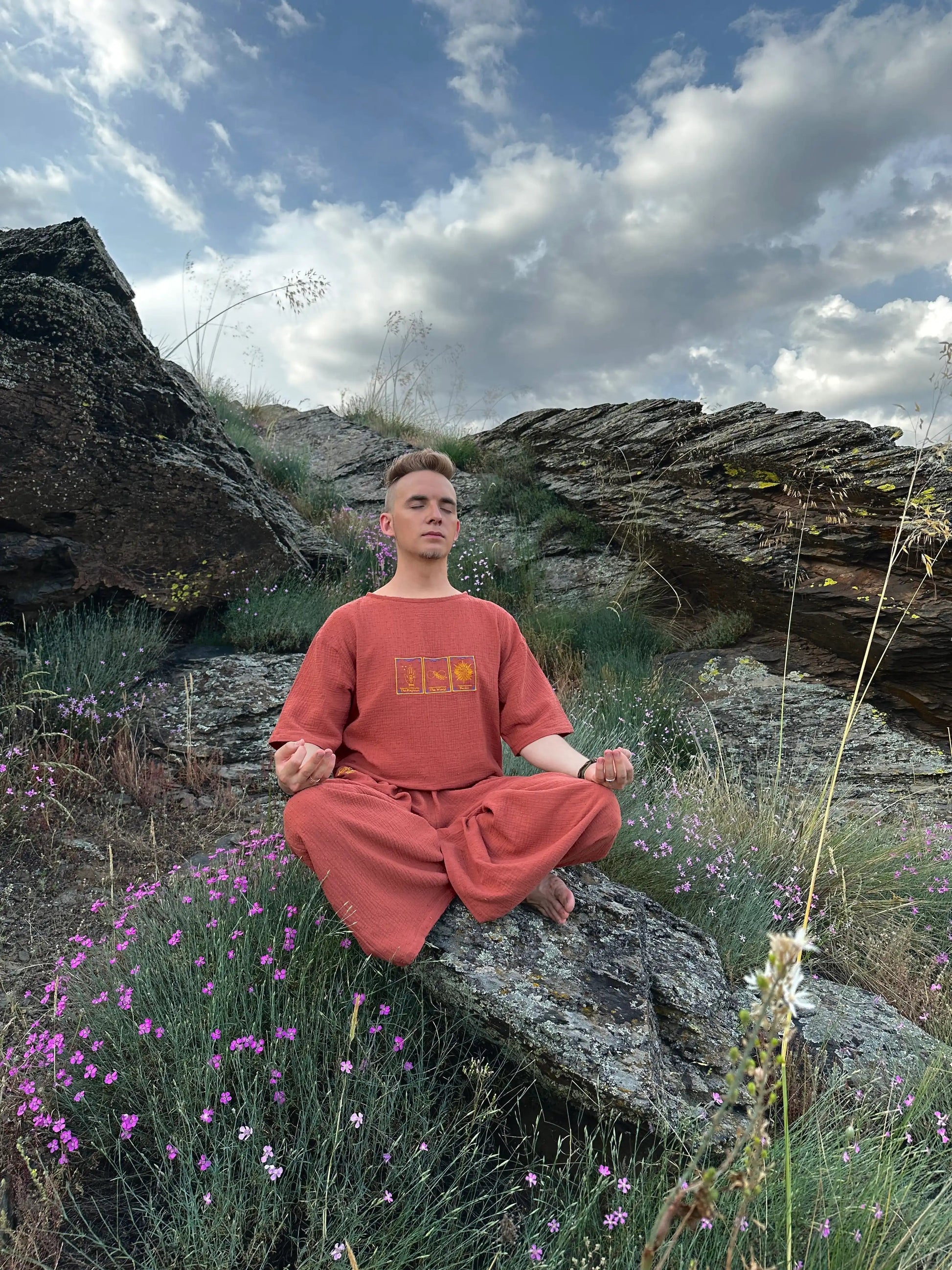 Person in orange outfit meditating on a rocky landscape with purple flowers and blue sky.