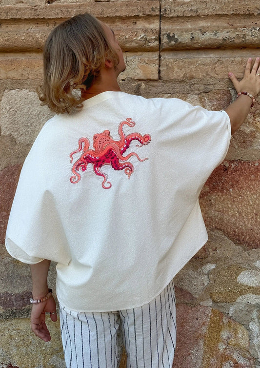 Person wearing a white shirt with a red octopus design, standing against a textured stone wall.