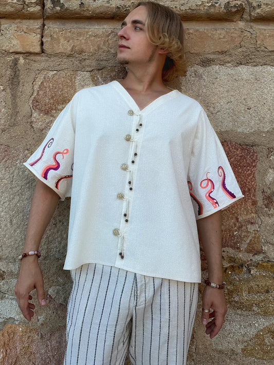 Person wearing a white embroidered shirt and striped pants standing against a stone wall.