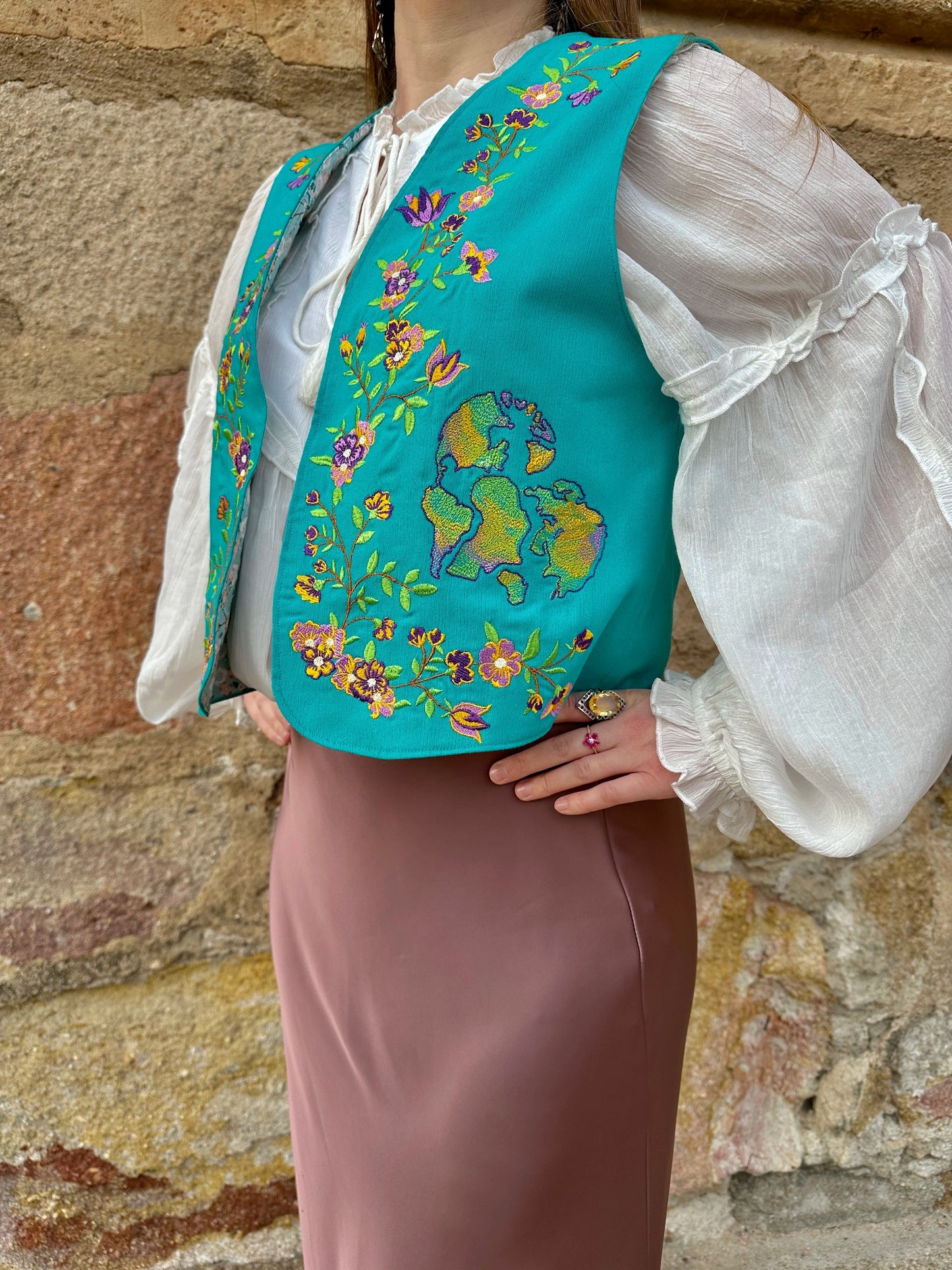 Person wearing a turquoise embroidered vest over a white blouse and brown skirt against a stone wall.