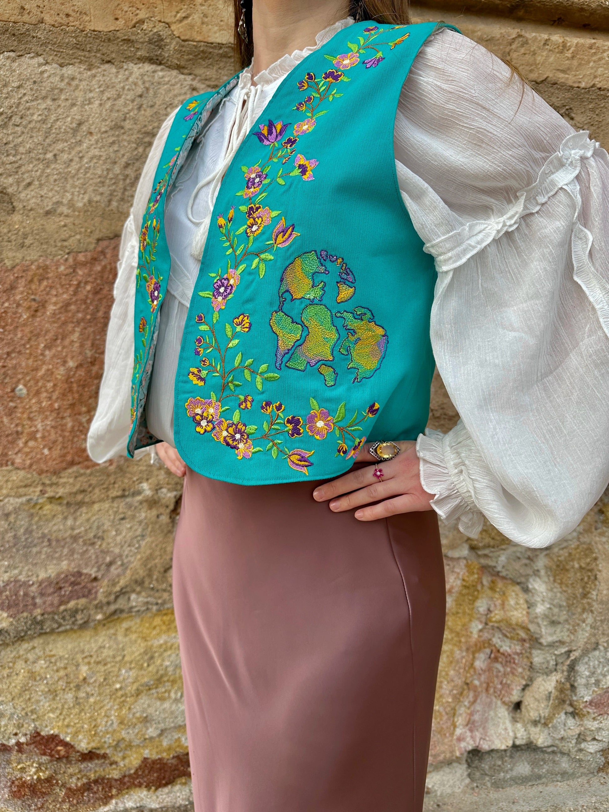 Person wearing a turquoise embroidered vest over a white blouse and brown skirt against a stone wall.