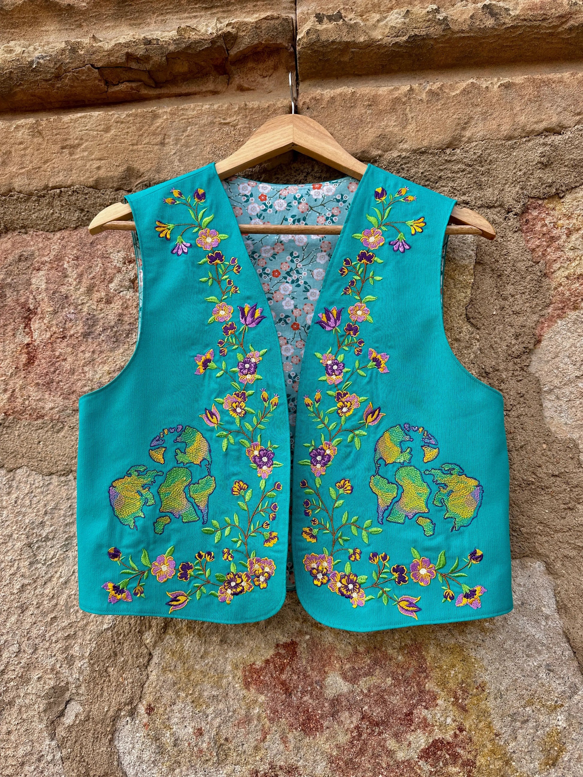 Turquoise embroidered vest on a wooden hanger against a stone wall.