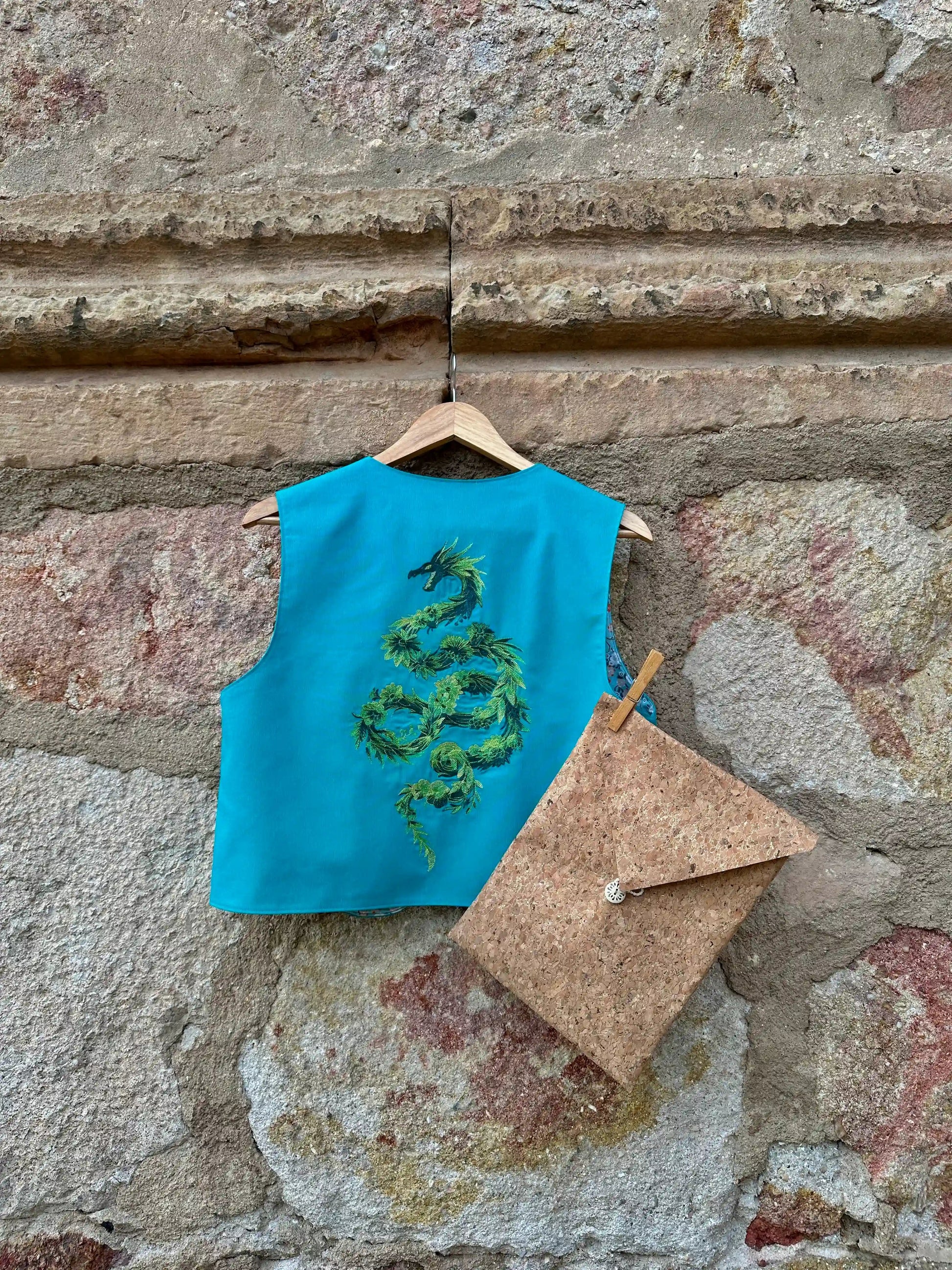 Turquoise top with green embroidery on a wooden hanger against a stone wall.