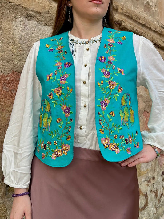 Woman wearing a traditional embroidered vest against a stone wall.