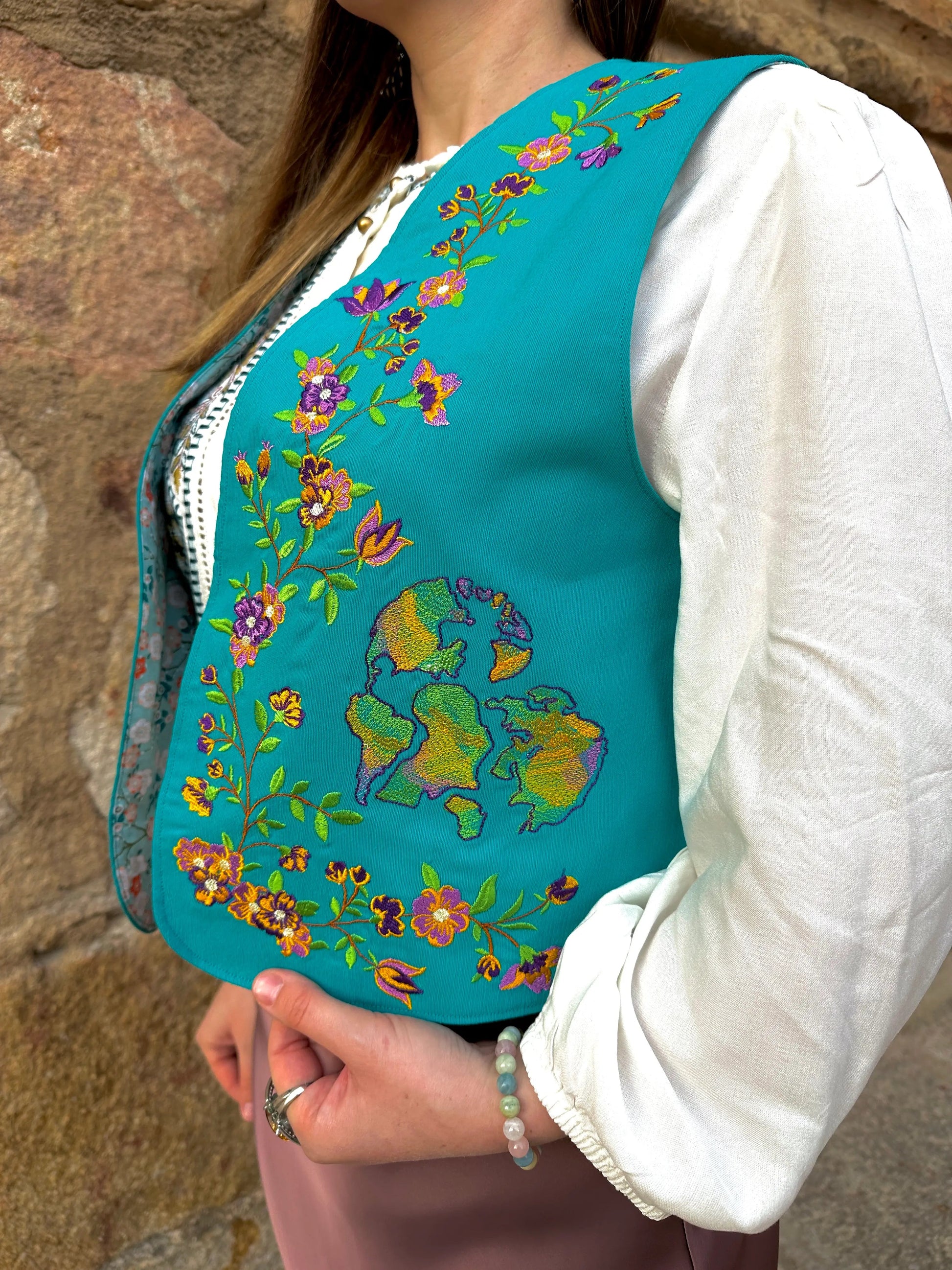 Teal embroidered vest over a white shirt with floral patterns, against a stone wall background.