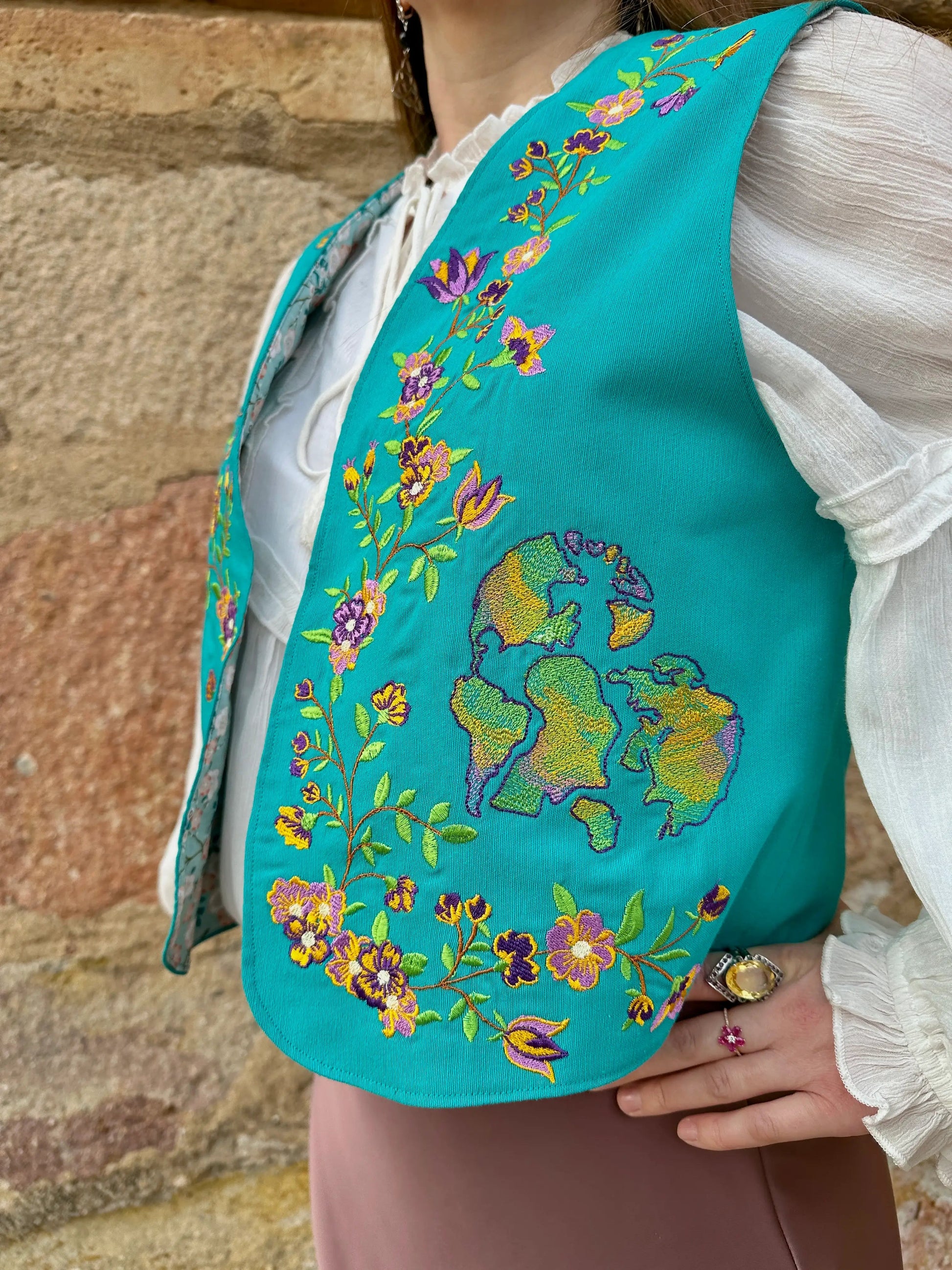 Teal embroidered vest with floral patterns worn by a person against a stone wall background