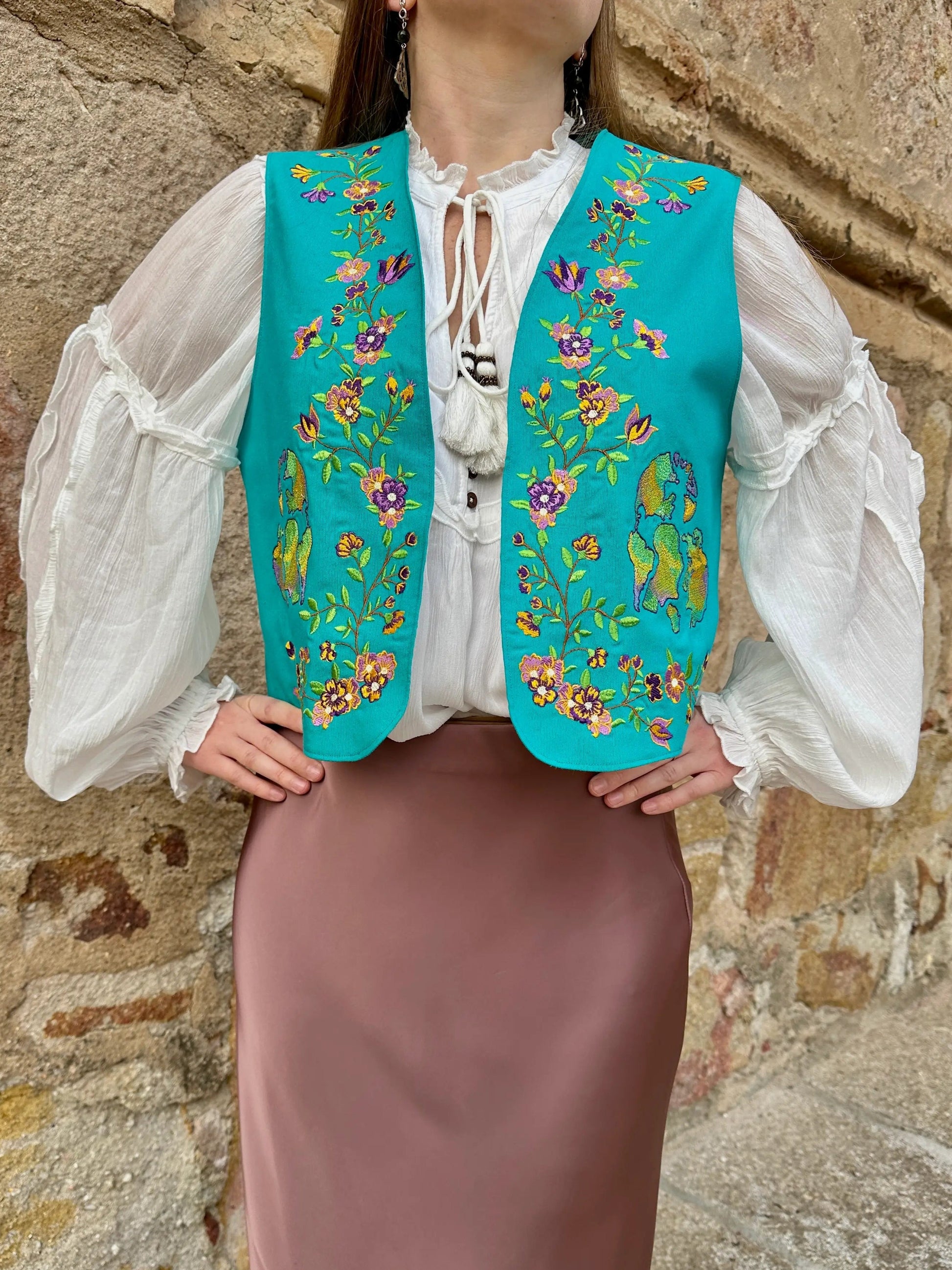 Person wearing a turquoise embroidered vest over a white blouse and brown skirt against a stone wall.