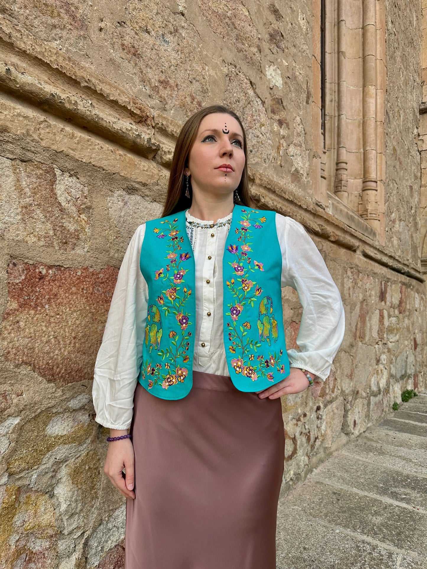 Woman wearing a traditional embroidered vest against a stone wall.