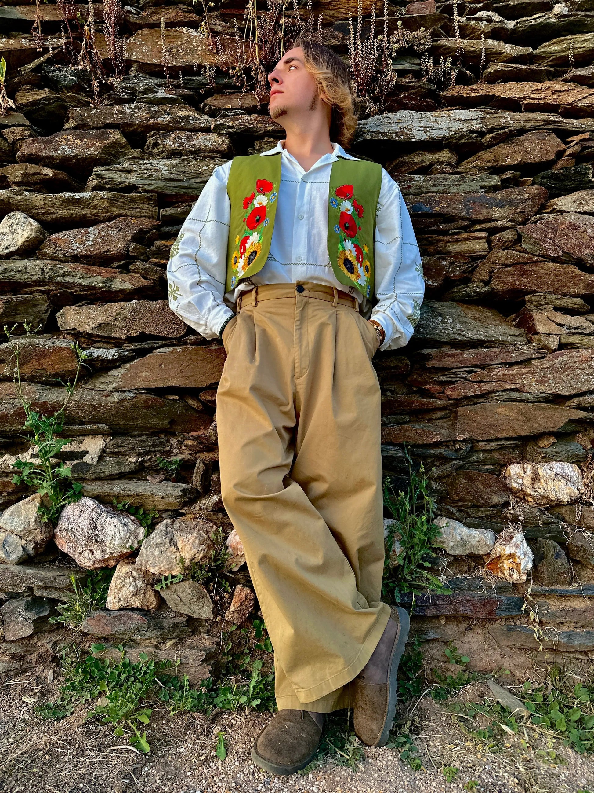 Person wearing a traditional outfit with a floral vest against a stone wall.