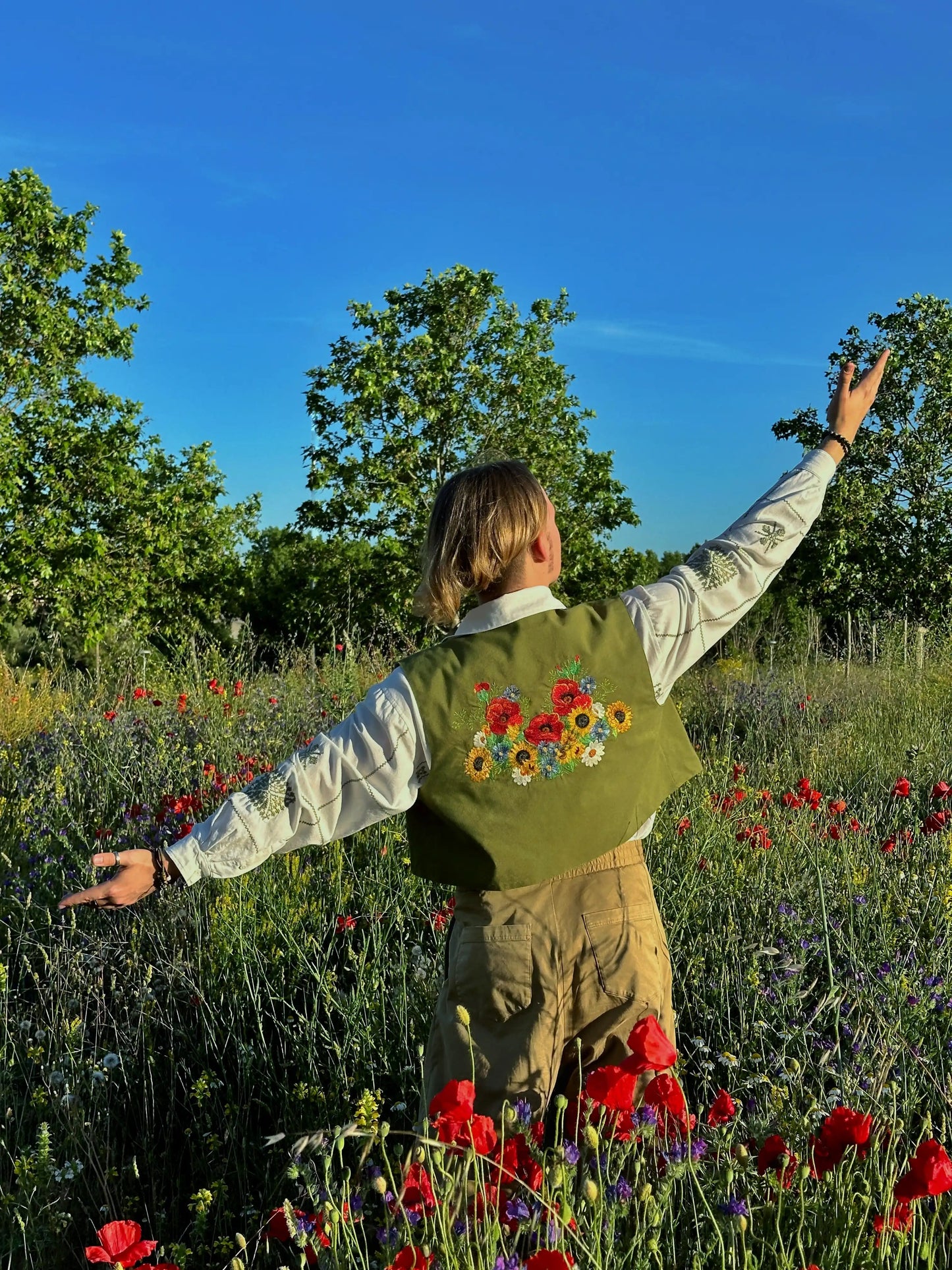 Person standing in a field of flowers with a green vest featuring embroidered flowers.