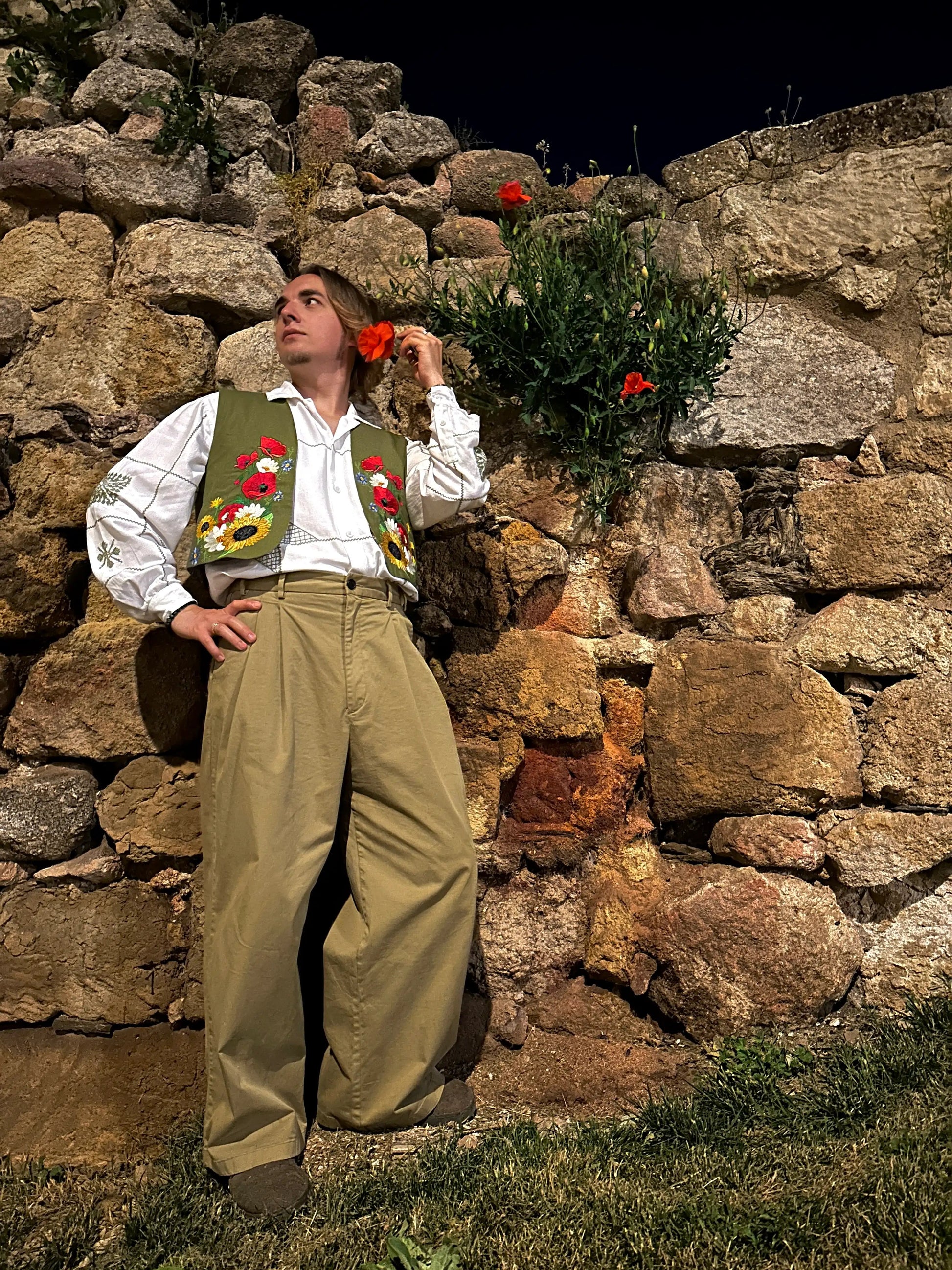 Person in traditional attire standing against a stone wall with flowers.