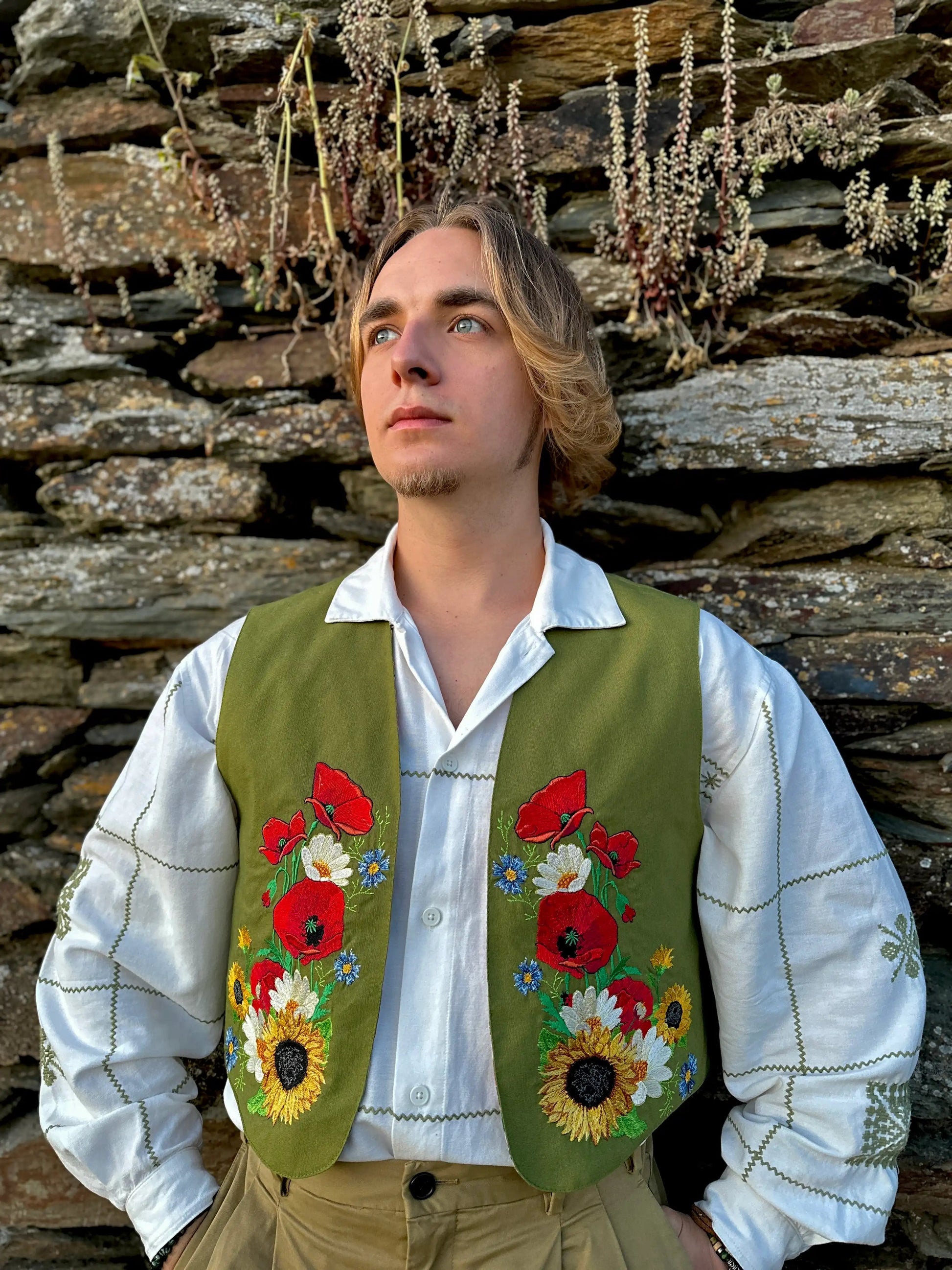 Man wearing a green embroidered vest with floral patterns against a stone wall.
