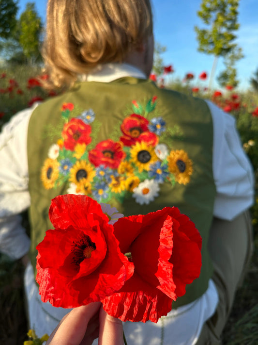 Person wearing a green vest with floral embroidery holding red poppies outdoors.