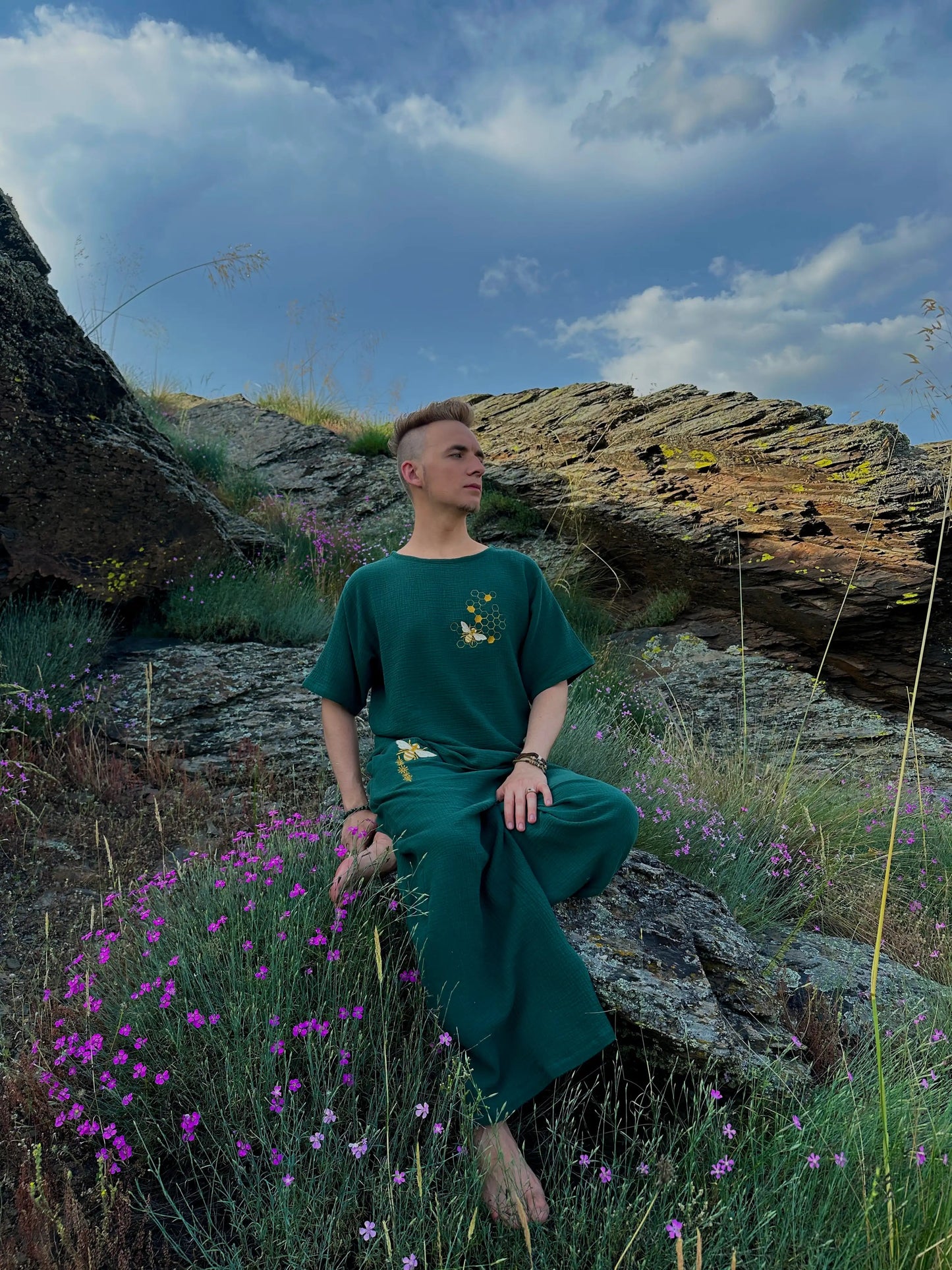 Person in a green outfit sitting on a rocky outcrop with purple flowers and a blue sky.