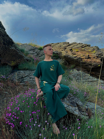 Person in a green outfit sitting on a rocky outcrop with purple flowers and a blue sky.