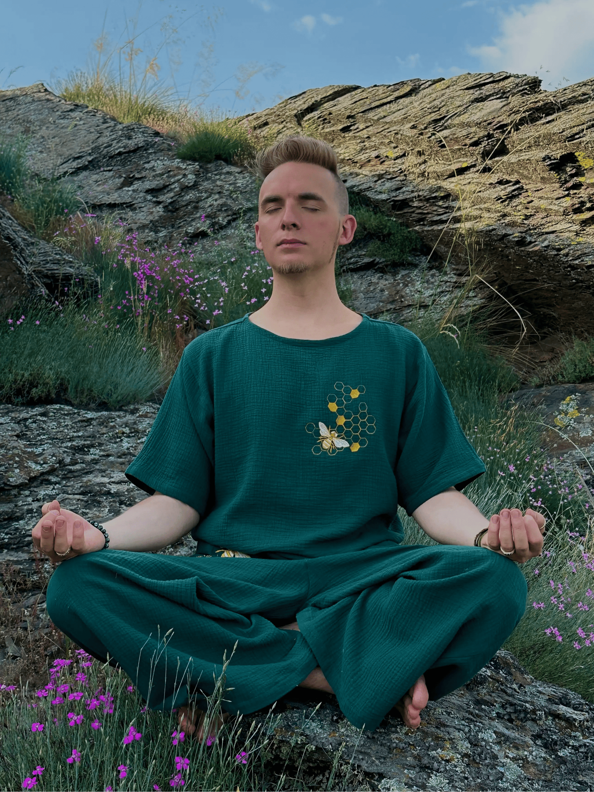 Person in a green outfit meditating on a rocky hillside with flowers and sky.