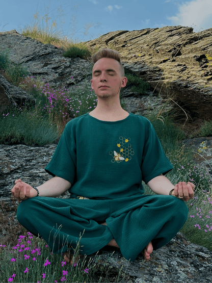 Person in a green outfit meditating on a rocky hillside with flowers and sky.