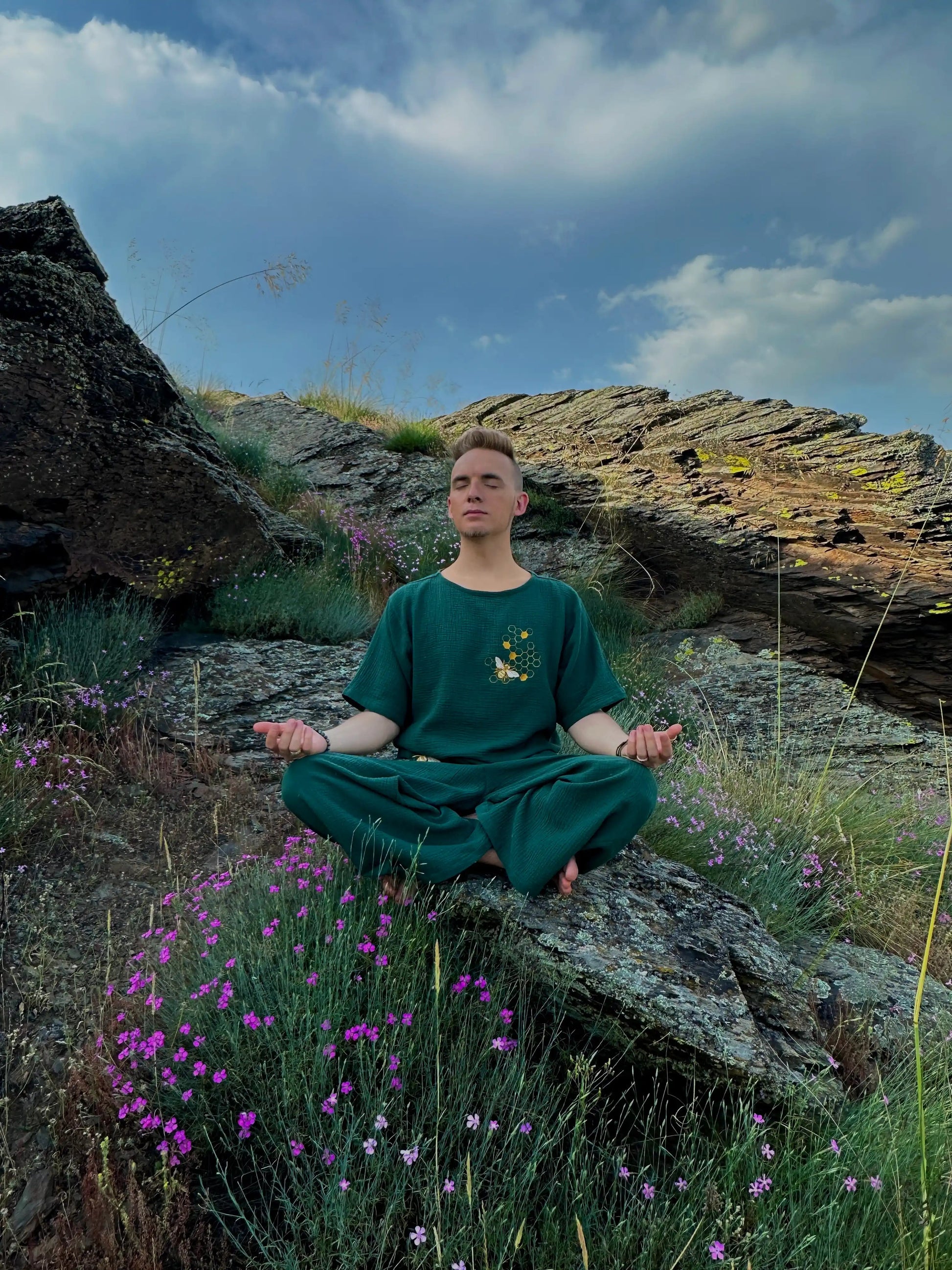 Person meditating on a rocky outcrop with purple flowers and a blue sky.