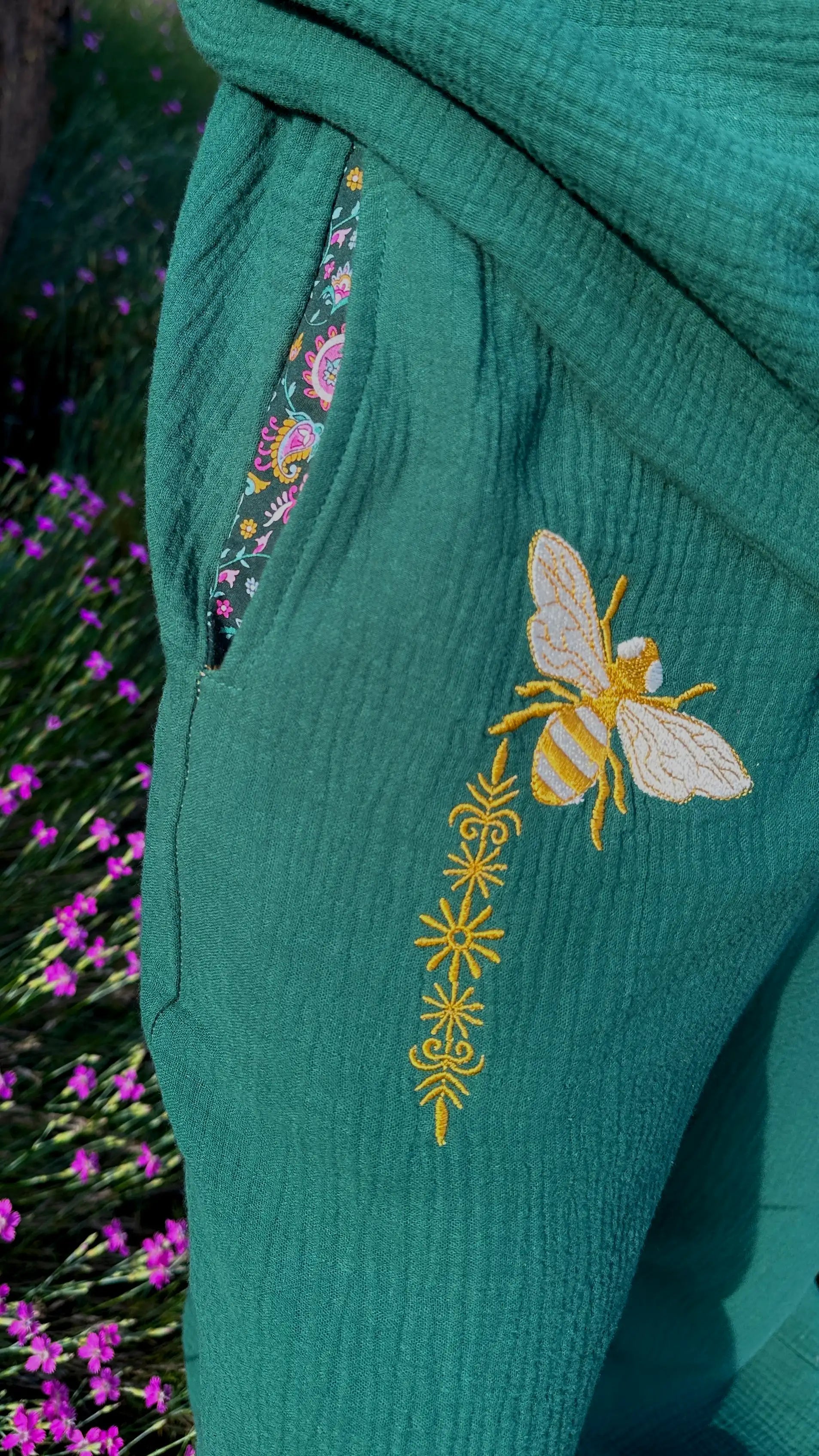 Green garment with embroidered bee design and floral trim against a blurred natural background