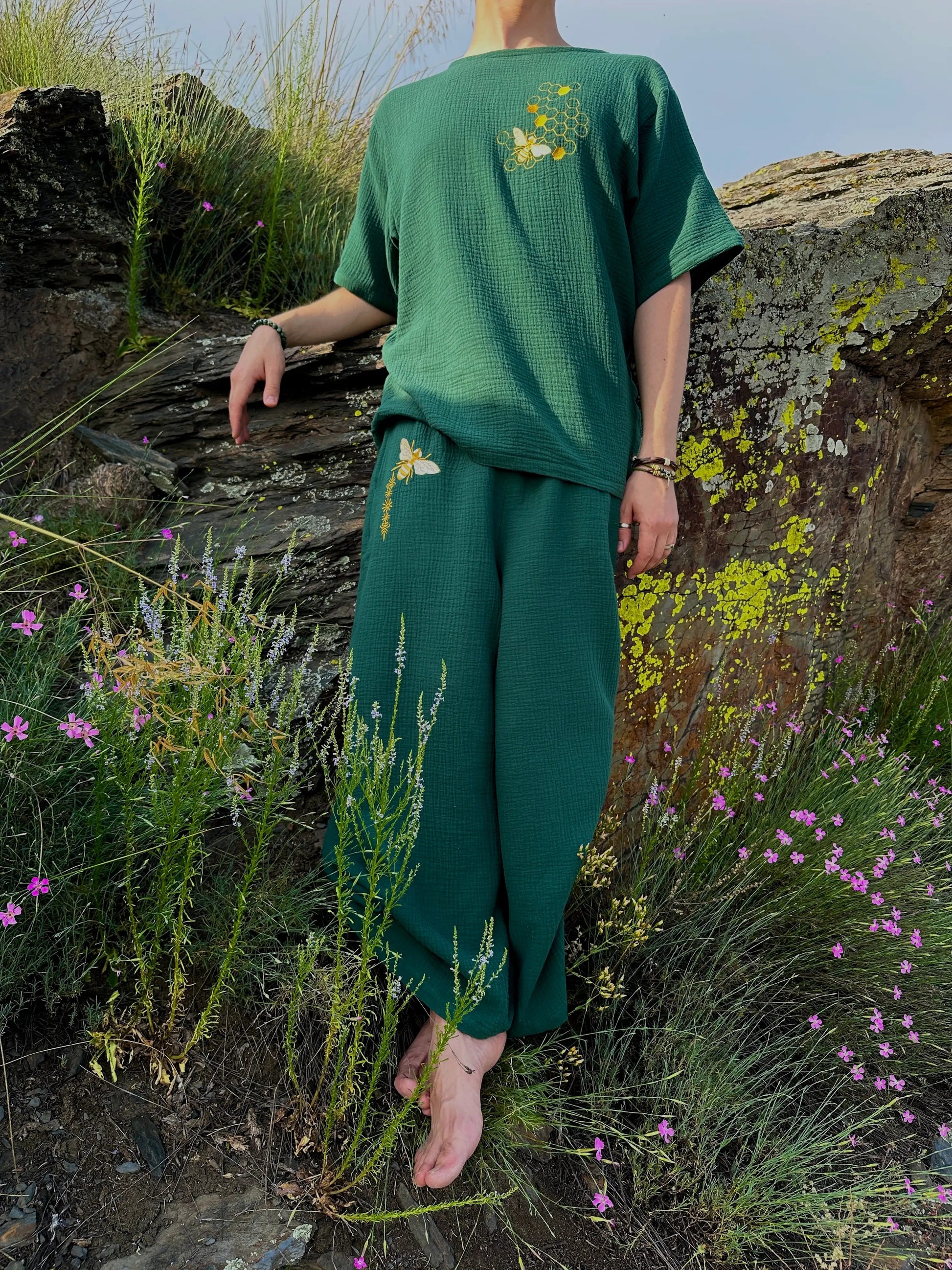 Person wearing a green outfit with floral patterns standing in a natural setting with rocks and flowers.