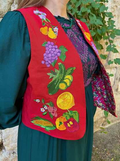 Person wearing a red embroidered vest with colorful vegetable patterns, standing against a stone wall.