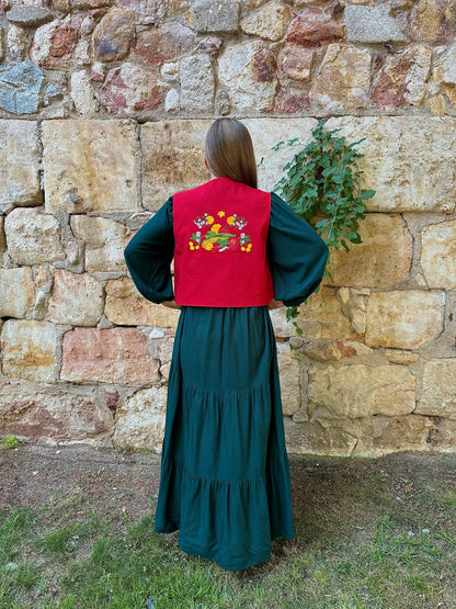 Person wearing a green dress and red vest with floral embroidery against a stone wall.