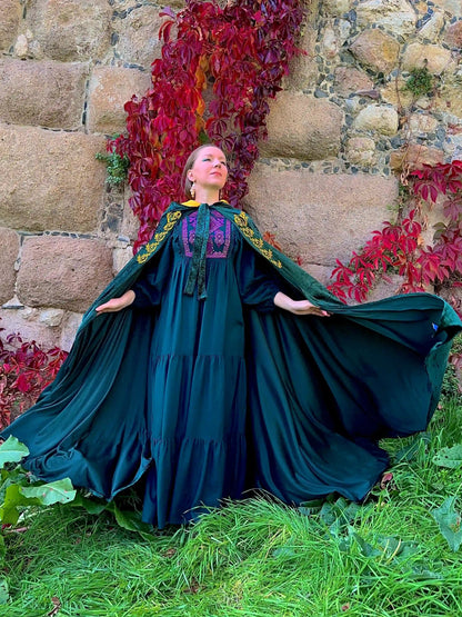 Woman in a long dark green dress with floral patterns standing in front of a stone wall and red foliage.