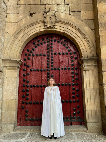 Person in a white cloak standing in front of a large red door with black metalwork on a stone building.