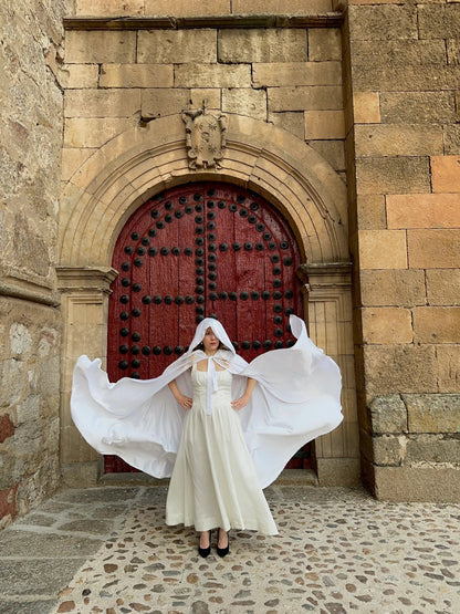 Woman in a white dress with a flowing cape standing in front of a large red door on a stone building.