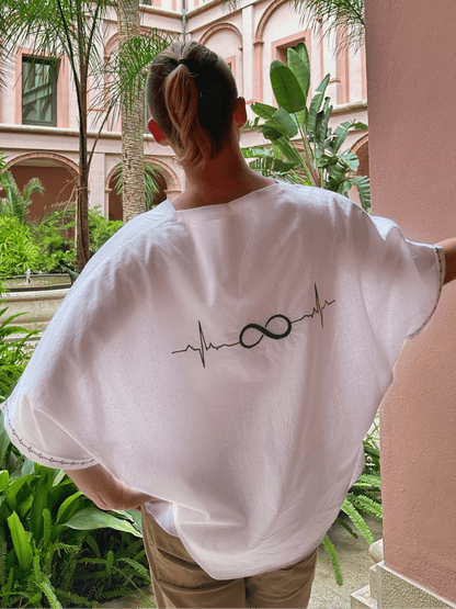 Person wearing a white shirt with a design, standing in a courtyard with pink walls and greenery.