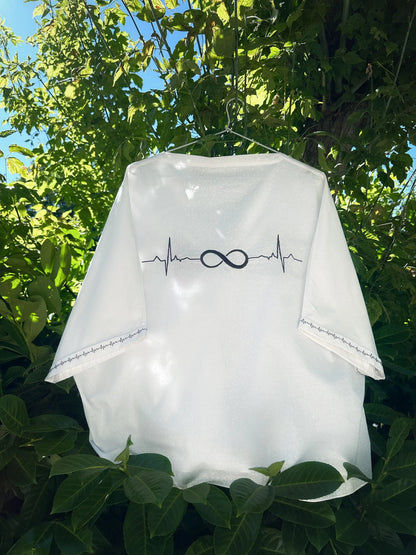 White baby romper with infinity symbol and heartbeat line design, hanging outdoors among green leaves.