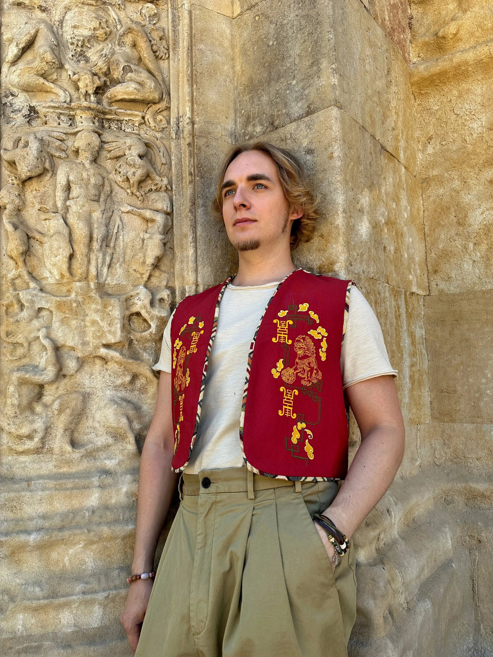 Person wearing a red vest with gold patterns against a stone wall with carvings