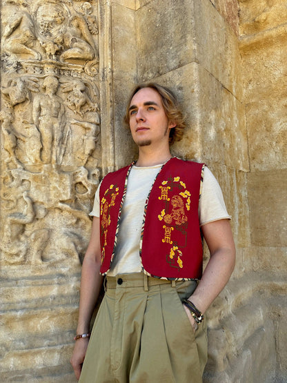 Person wearing a red vest with gold patterns against a stone wall with carvings