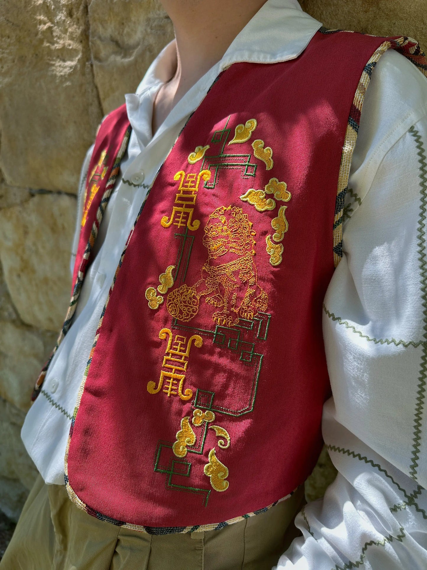 Person wearing a red embroidered vest over a white shirt against a stone wall.