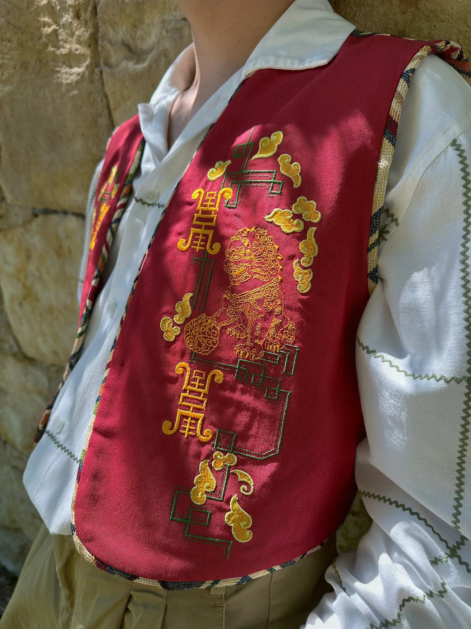 Person wearing a red embroidered vest over a white shirt against a stone wall.