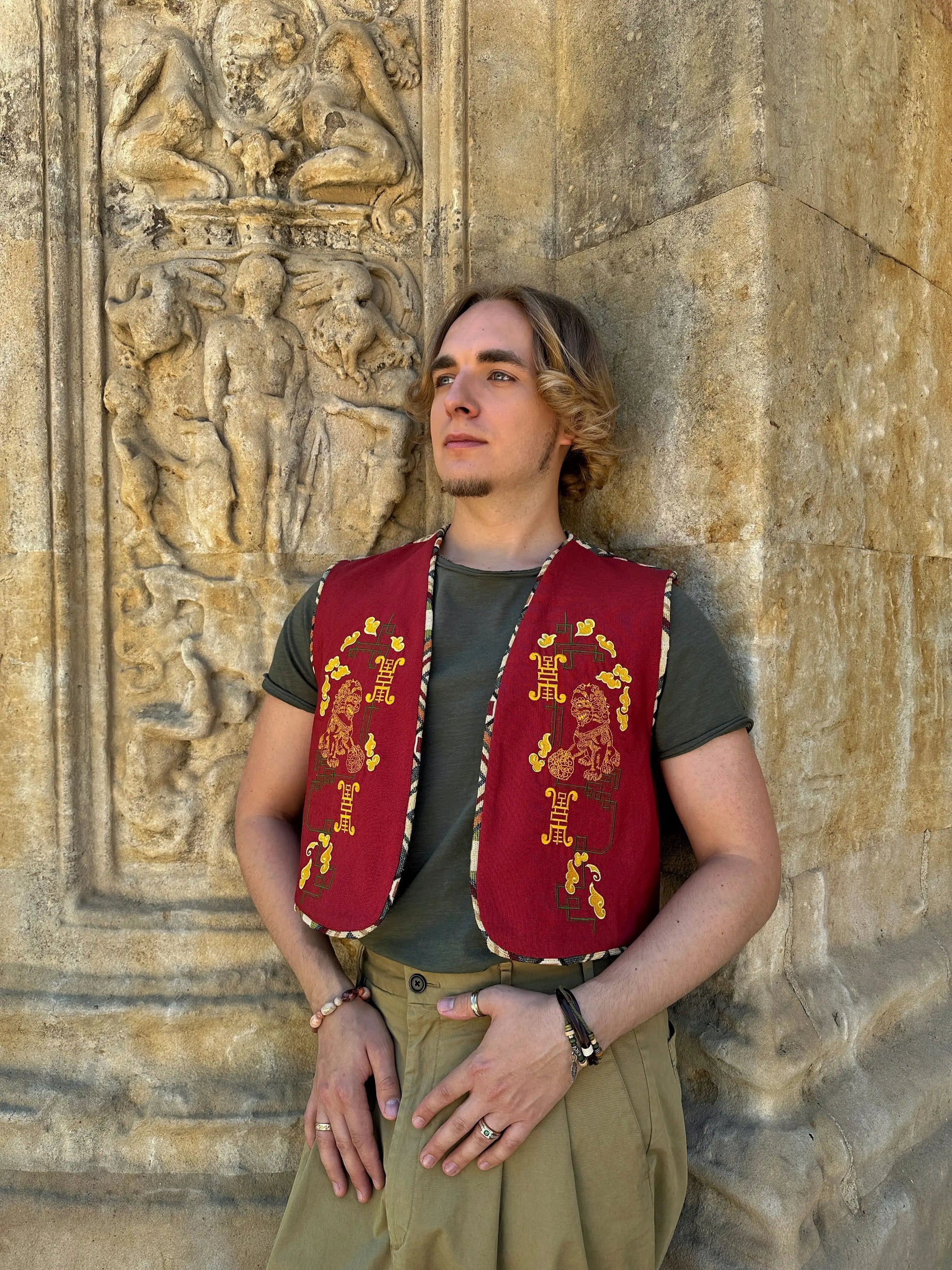 Person wearing a red vest with gold embroidery against a stone wall with carvings