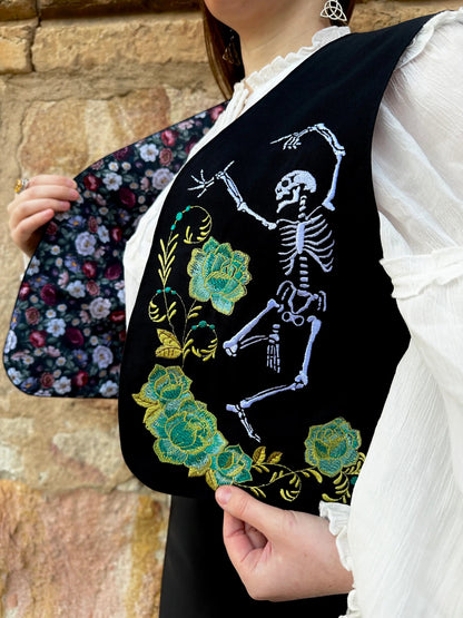 Person holding a black embroidered vest with a skeleton and floral design against a stone wall.