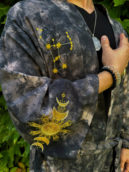 Person wearing a dark, tie-dye jacket with embroidered celestial designs on a green leafy background.