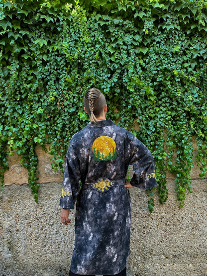Person wearing a black tie-dye robe with a sun design, standing against a wall covered in green ivy.